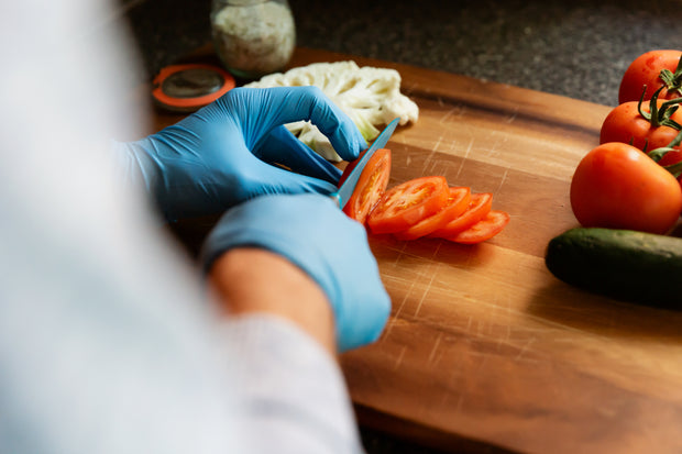 Lebensmittelzubereitung: Person mit blauen Einweghandschuhen schneidet Tomaten auf einem Schneidebrett, frische Zutaten im Hintergrund